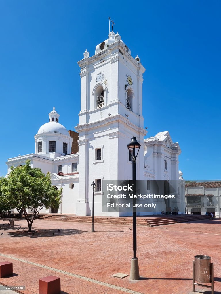 White colonial-style cathedral with a prominent clock tower and dome, set against a clear blue sky in a red-brick plaza with street lamps and a few trees in Santa Marta, Colombia.